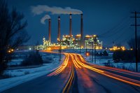 Industrial power plant with illuminated smokestacks at night, light trails from traffic on snowy road leading to factory complex against dark blue winter sky.