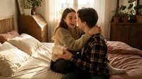 Young couple sharing intimate moment laughing together on bed in cozy bedroom with warm natural light streaming through window creating romantic atmosphere.