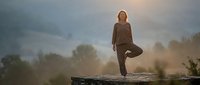 Mature woman practicing yoga tree pose on wooden platform at sunset with peaceful mountain landscape background for wellness and mindfulness concepts.