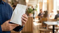 Man in dark blue shirt holding white napkin or paper towel in restaurant or cafe setting with blurred background and other guests dining.