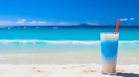 Tropical blue cocktail with red straw on pristine white sand beach with turquoise ocean waves and clear sky for summer vacation and relaxation concepts.