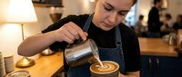 Young female barista creating latte art in coffee cup at cafe counter, pouring steamed milk from metal pitcher into cappuccino with focused concentration.