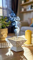 Elegant ceramic incense burner with rising smoke on wooden table, alongside yellow candle and houseplant, creating peaceful home atmosphere in soft natural light.