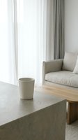 Minimalist ceramic cup on concrete table in bright living room with beige sofa, wooden furniture, and sheer curtains. Interior design and home decor.