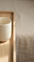 Ribbed ceramic mug with cream-colored glaze sitting on wooden tray against neutral beige wall, minimalist home decor and tableware styling.