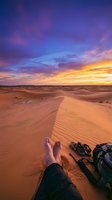 Traveler sitting on sand dunes at sunset with boots and backpack, enjoying dramatic sky with golden and purple hues over desert landscape. Perfect for travel and adventure content.