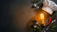 Professional chef hat with vintage Edison bulb and culinary utensils on dark background creating warm atmospheric lighting for restaurant kitchen concept.