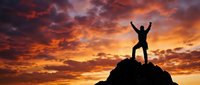 Silhouette of triumphant person with raised arms standing on mountain peak against dramatic orange and red sunset sky. Success, achievement, victory concept.