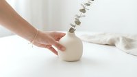 Caucasian hand arranging dried eucalyptus stems in white ceramic vase on neutral surface for home decor, interior styling, and botanical arrangement inspiration.