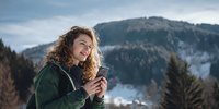 Young woman with curly hair in green jacket using smartphone against mountain landscape, enjoying nature during winter hiking adventure.