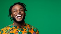 Young African American man with dreadlocks and beard wearing bright orange tropical print shirt laughing joyfully against green background, expressing genuine happiness and positive emotions.