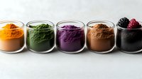 Colorful superfood powder collection in glass jars including turmeric, spirulina, acai, cacao and activated charcoal with fresh berries on white background.