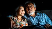 Senior Asian couple relaxing together in dark room, elderly woman resting head on husband's shoulder, intimate emotional moment captured in dramatic lighting.