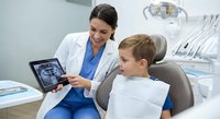 Female dentist showing dental X-ray results to young blonde boy patient in modern clinic chair. Professional healthcare consultation and pediatric dentistry.