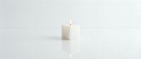 White pillar candle with a glowing flame on a reflective surface, soft light and minimalist style for spa, wellness, and relaxation concepts.
