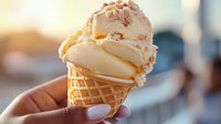 Creamy vanilla ice cream scoops in waffle cone topped with crushed nuts, held by feminine hand with neutral manicure against soft blurred background.