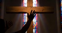 Person reaching toward wooden cross in church with colorful stained glass windows creating spiritual atmosphere for worship and prayer.