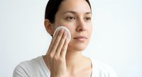 Young Hispanic woman applying skincare product with cotton pad on white background for beauty and wellness routine.