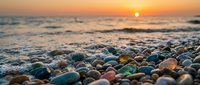 Colorful smooth pebbles scattered on beach shore during golden sunset over ocean waves. Natural coastal landscape with warm lighting for relaxation themes.