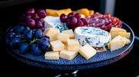 Gourmet cheese board with blue cheese, cheddar cubes, red and purple grapes, and cured meats arranged on dark textured plate, elegant appetizer presentation.