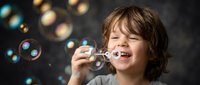 Happy young boy with brown hair blowing soap bubbles, creating magical floating spheres against dark background for childhood joy and play concepts.