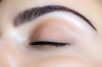 Close-up view of closed eye with professionally groomed eyebrow, black eyeliner, and neutral eyeshadow showing detailed makeup application technique.