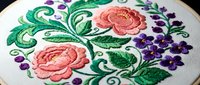 Embroidered floral design with pink roses and purple flowers in decorative hoop, traditional needlework craft on white fabric background.