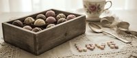 Assorted chocolate truffles in a rustic wooden box on a lace tablecloth, with a vintage teacup and spoon in soft, warm tones.
