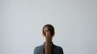 Woman with long brown hair in ponytail wearing dark jacket, rear view against neutral background, suitable for business, corporate, and professional design projects.