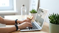 Female hands typing on laptop computer at modern workspace with plants and water bottle creating productive home office environment for remote work.
