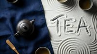 Zen tea ceremony flat lay with clay teapot, cups, and TEA lettering drawn in white sand on blue linen fabric, top view.