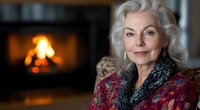 Elegant senior Caucasian woman with silver hair wearing colorful patterned clothing sits by warm fireplace in cozy home interior for lifestyle content.