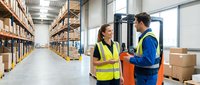 Two warehouse workers in safety vests discussing logistics near forklift in modern distribution center with organized shelving and inventory management.