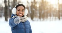 Happy African American woman in winter coat and earmuffs pointing at camera in snowy forest with warm sunlight creating magical atmosphere.