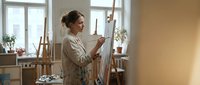 Young Caucasian woman artist painting on canvas in bright art studio workspace with easel and natural window light for creative projects.