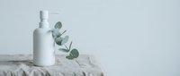 White cosmetic bottle with spray pump and fresh eucalyptus branches on textured stone surface, skincare and wellness product display.