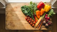 Fresh organic vegetables arranged on rustic wooden cutting board including kale, bell peppers, carrots, radishes and cherry tomatoes for healthy cooking.