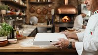 Mature male chef in white chef's coat using tablet in rustic kitchen with wood-fired oven, copper cookware and herbs, for culinary business and restaurant management concepts.