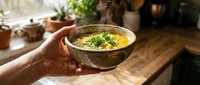 Hands holding ceramic bowl of creamy soup garnished with fresh herbs on rustic wooden table in cozy kitchen setting for comfort food and homemade cooking concepts.