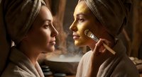 Two young Caucasian women in towels at spa, one applying gold face mask with brush, warm luxury wellness atmosphere.