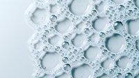 Macro view of soap bubbles forming delicate foam pattern in light blue and white tones, creating abstract geometric circles and organic shapes with transparent texture.