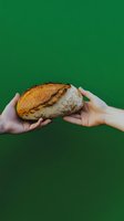 Hands passing freshly baked artisanal sourdough bread loaf on green background, symbolizing sharing and community spirit in food culture.