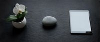 Minimalist dark slate desk with white orchid in pot, zen stone, and tablet with blank screen for mockup display.