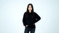 Young Asian woman in black sweater and jeans smiling confidently with hands in pockets against white background for lifestyle and fashion content.