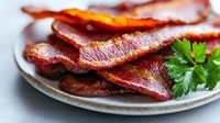 Crispy fried bacon strips served on ceramic plate with fresh parsley garnish, close-up view showing texture and caramelization of cooked meat.