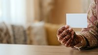 Woman with henna-decorated hands holding blank white card at wooden table, suitable for greeting cards, invitations, and cultural event marketing materials.