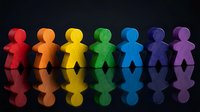 Seven colorful paper cutout figures in rainbow colors standing in row with reflections on dark surface representing diversity and inclusion.