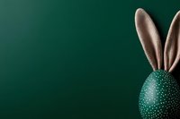 Green Easter egg decorated with white polka dots and bunny ears peeking from corner against dark emerald background, minimal holiday composition.