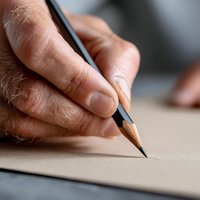 Close-up of mature hand holding pencil writing on paper, showing detailed skin texture and focused grip for creative work and artistic expression.