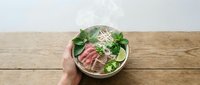 Unrecognizable person holding bowl with fresh salad, sliced meat, noodles and greens on wooden table for healthy eating and nutrition content.